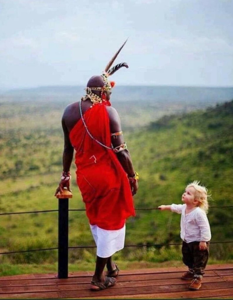 image shows a black man in natice dress, appears to be African, and a very small blond girl child on a bridge overlooking a hillside forest